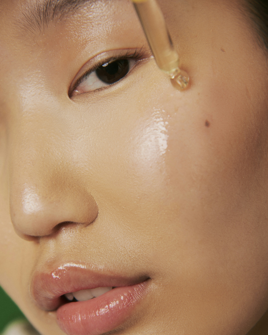 Close-up of glowing, hydrated skin on a woman’s face as a glass dropper applies a clear facial oil or serum to her cheek, with natural light highlighting smooth texture and a soft, dewy finish.